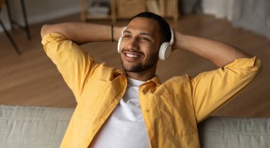 Handsome young black guy in headphones relaxing on couch, listening to music at home, banner design. Millennial African American man enjoying audio book in headset, resting indoors