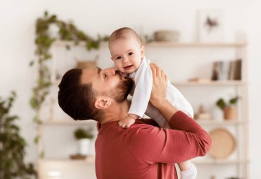 Babalar sever. Sevimli, genç bir babanın küçük kızını öpmesi ve kucağına alıp kucağına alması kucağında kucağında yeni yürümeye başlayan bebekle evde durması. Ebeveynlik ve Babalık Keyfi