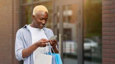 Kişisel indirim, alışveriş ve tüketim. Elinde paketler olan gülümseyen Afro-Amerikan adam, butiğin yanında akıllı telefondan yazı yazıyor.