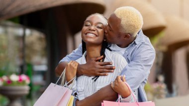 Alışverişte sarılmalar. Neşeli Afro-Amerikalı adam dükkanların yanında renkli çantalı kızı öpüyor, güneş parlıyor, boş yer var.