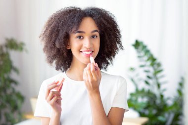 Nemlendirici dudaklar ve hassas derinin soyulmasını önler. Milenyum Afrikalı Amerikalı kadın çıplak makyaj yapıyor. Profesyonel makyaj malzemesi kullanıyor. Oturma odasında ruj sürüyor.