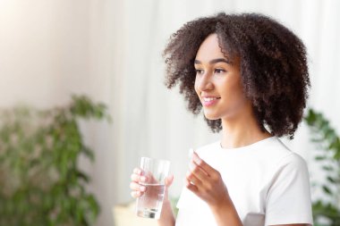 Diyet, beslenme, sağlıklı beslenme, modern yaşam tarzı, mineraller ve vitaminler. Neşeli genç Afro-Amerikalı kadın, oturma odasında, yan tarafta, fotokopi odasında bir bardak suyla hap ve içecek alıyor.