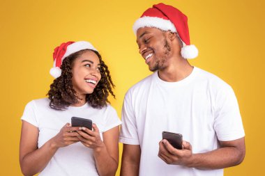 Noel 'i kutlayan akıllı telefonlar kullanan Afrikalı Amerikalı çift Sarı Stüdyo Arkaplan' ın başında dikilen kameraya gülümsüyor. Mobil Uygulama Xmas Reklamı. Panorama