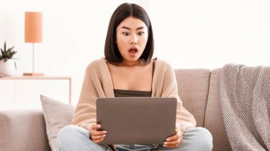 Omg, son dakika haberleri. Şaşırmış Asyalı kadın dizüstü bilgisayarda beklenmedik bir şey izliyor, evdeki kanepede oturuyor, boşluğu kopyalıyor. Ağzı açık genç bir bayanın portresi
