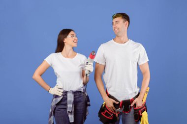 Matkap ve güvenlik gözlüklü genç bir çift onarımlara başlamaya hazır, milenyum eşleri mavi stüdyo arka planında inşaat aletleriyle ayakta duruyor ve birbirlerine gülümsüyor, uzay kopyaları yapıyorlar.