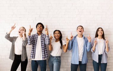 Mutlu genç arkadaşlar beyaz tuğla duvarın üzerinde boş alanı gösteriyorlar, sıra sıra duruyorlar.