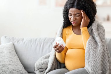 Baş ağrısından muzdarip hamile bir zenci kadın. Ağrı kesici alıyor. Oturma odasının içinde. Hasta Afro-Amerikalı, elinde haplar olan bir kadın bekliyor. Evdeki koltukta oturuyor, fotokopi çekiyor.