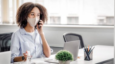 Yöneticiler karantinadan sonra müşterilerle çalışırlar. Koruyucu maskeli Afrikalı Amerikalı kadın telefonda konuşuyor, iş yerinde dizüstü bilgisayarla oturuyor, modern ofis içinde antiseptik, gündüz vakti serbest alan