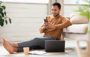 Portrait of young man holding using cell phone sitting on floor leaning on couch in living room. Cool guy browsing internet, surfing web watching video, using application and pc, full body length