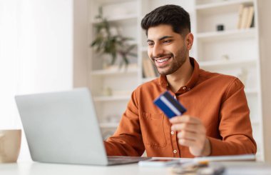 Hızlı Çevrimiçi Alışveriş. Elinde banka kartı tutan ve bilgisayarını kullanan gülümseyen genç bir adamın portresi oturma odasındaki masada oturarak finansal işlem yapmak, bedava fotokopi alanı.