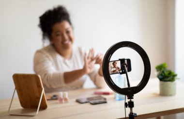 Cheerful pretty curly young chubby hispanic lady beauty blogger sitting at desk full of various makeup products, recording video on smartphone, showing different nail poloshers, recommending cosmetics