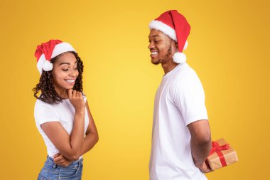 Noel için sürpriz. Siyah Erkek Arkadaş, Arkasında Paketlenmiş Hediye Kutusu Tutuyor Kız Arkadaşını Sarı Stüdyo Arkasında Dururken, Noel Baba Şapkaları Giyerken Kutluyor. Noel 'i Kutlayan Afrikalı Çift