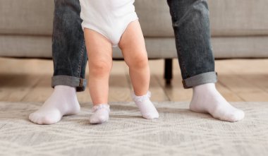 Baba, bebeğinin ilk adımlarını atmasına evde birlikte yürümeyi öğrenmesine yardım ediyor. Babalar ve Çocuk Bacakları. Babalık ve Sevimli Aile Anları kavramı. Kırpılmış