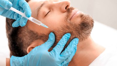 Estetik kliniğinde güzellik iğnesi yaptıran sakallı adamın yakın çekimi. Estetik cerrah yakışıklı erkek yanaklarına yaşlanma önleyici enjeksiyon yapıyor. Erkek kozmetolojisi, estetik tıp konsepti