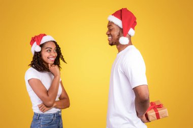 Noel hediyeleri. Siyah adam paketlenmiş Xmas Hediye Kutusu Sarı Stüdyo Arkaplanında Bekleyen ve Noel Baba Şapkaları Giyen Kız Arkadaşınızı Kutluyor. Yılbaşı sürprizi hazırlanıyor, Kış Tatili Kutlaması