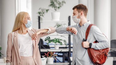 Coronavirus salgını sırasında meslektaşlarımızla modern selamlaşmalar. Milenyum adamı ve koruyucu maskeli kadın sabah işe geldiler ve dirsekleriyle dokunarak ofis içi boşluğa merhaba dediler.