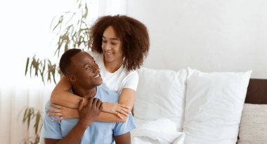 Çekici erkek ve kadın yatak odasında, aşkta ve mutlulukta eğleniyor. Neşeli genç Afro-Amerikalı kadın, kocasının arkasından sarılıyor. Sabahları rahat bir şekilde yatağında oturuyor.