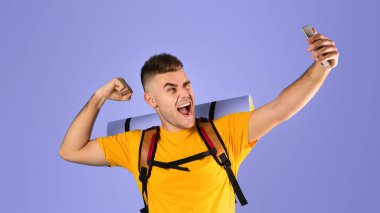 Turist ekipmanlarıyla havalı genç adam mavi stüdyo geçmişi üzerinden akıllı telefondan selfie çekiyor. Komik bir milenyum yürüyüşçüsü cep telefonuyla komik bir fotoğraf çekiyor. Seyahat macera konsepti
