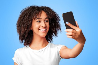 Modern teknoloji, mobil uygulama ve moda blogu için selfie. Gülümseyen Afro-Amerikan milenyum kadını, mor arka planda izole edilmiş akıllı telefondan fotoğraf çekiyor.