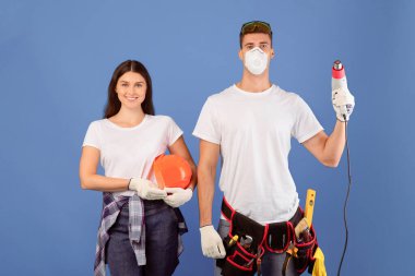 Ev tamiri. Sert şapkalı ve koruyucu maskeli genç çift mavi arka planda inşaat aletleri olan, milenyum adamı kameraya ve kasklı kadına sondaj yapmayı hedefleyen, serbest alan.