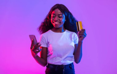 Cep telefonu ve kredi kartı tutan neşeli Afrikalı Amerikalı genç kadın, online ticaret veya kumar, bankacılık veya alışveriş mobil uygulaması kullanarak, neon ışıkta nakit para alıyor