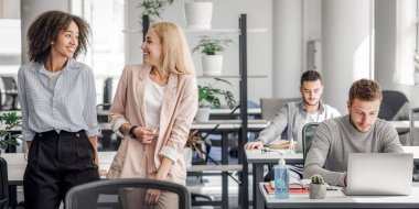 İşe dönüyorum, yeni normal ve tecritten sonra iletişim kuruyorum. Mutlu Avrupalı ve Afro-Amerikan kadınları modern ofisin içinde iş arkadaşlarıyla salgın sırasında iş yerlerinde dedikodu yapıyorlar.