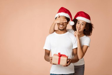 Romantik Noel ve Yeni Yıl birlikte. Noel Baba şapkalı neşeli Afro-Amerikalı kadın elinde sessizlik işareti gösteriyor ve gözlerini kapatıyor açık arkaplanda hediye kutusu olan kocaya, boş alana, stüdyo çekimine.