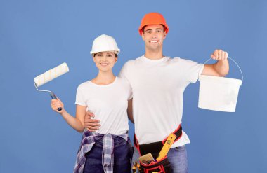 Ev tadilatı. Mavi arka planda boyama aletleri olan gülümseyen genç bir çiftin portresi. Apartman onarımına hazır, elinde boya ve tekerlekli fırçayla kovayı tutan neşeli bir adam ve kadın.