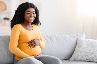 Gülümseyen Afro-Amerikan kadın kucağında bebek, koca göbeğine sarılmış evde kanepede otururken. Neşeli siyah hamile kadın karnını kucaklıyor ve kameraya gülümsüyor, anne olmaktan mutlu.