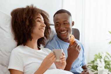 Sabahları yatakta kahvaltı ve karantina sırasında keyifli bir ruh hali. Gülümseyen Afrikalı genç Amerikalı karı koca ellerinde fincanlar, sıcak kahve, kruvasan yiyip, yatak odasında sohbet ediyorlar.