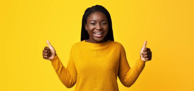 Hoşuma gitti. Parlak kazaklı neşeli Afrikalı kadın kamerada başparmaklarını gösteriyor, olumlu siyah kadın bir şey öneriyor, onaylama işareti yapıyor, sarı arka planda duruyor.