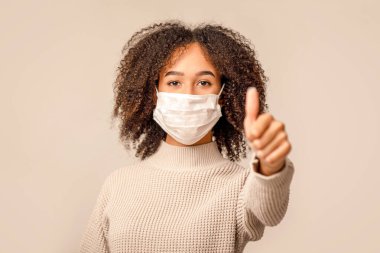 Güzel, onaylayın. Milenyum dostu Afro-Amerikan kadın koruyucu maskeli. Başparmaklarını göstererek Coronavirus salgınıyla mücadele ediyor. Krem rengi arka planda izole edilmiş.
