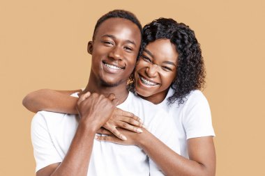 Afrikalı Amerikalı genç adam ve kadının sarı stüdyo arka planında sarılışının portresi. Mutlu siyahi aşıklar sarılıyor, duygusal bayan neşeli erkek arkadaşına arkadan sarılıyor, yakın plan.
