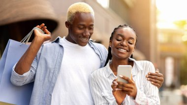 Akıllı telefondan reklam ve indirimler yolluyorlar. Alışveriş çantalı mutlu Afro-Amerikan çifti butiğin yanındaki telefona bakıyorlar.