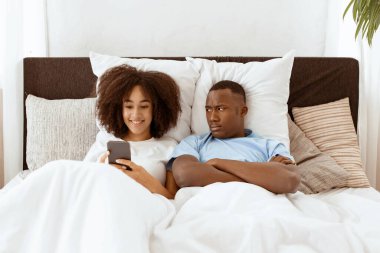 Koca internette ve sosyal ağlarda karısına olan bağımlılığından rahatsız oluyor. Huysuz, kızgın, Afro-Amerikalı bir adam yatakta gülümseyen bir kadının akıllı telefonuna bakıyor.