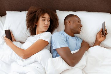Kıskançlık, sorunlar, internette ve sosyal ağlarda hile yapmak. Milenyum şüphelisi Afro-Amerikan kadın yatak odasında gülümseyen bir erkeğin telefonuna bakıyor. İç mekan, üst manzara, serbest alan.