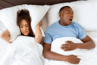 Aile sorunları, apne ve uykusuzluk çeken bir eş. Genç, yorgun Afrikalı Amerikalı adam uyuyor ve horluyor, uykusuz kadın kulaklarını yastıkla kapatıyor.