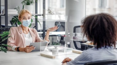 Coronavirus salgını ve sağlık korunması sırasında modern insan kaynakları ve röportaj. Koruyucu maskeli müdür, modern ofis içi koruyucu cam aracılığıyla Afrikalı Amerikalı kadınlara sorular soruyor.