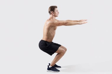 Gövdesi çıplak, atletik bir adamın hafif stüdyo arkaplanı üzerinde aerobik egzersizleri yaparken yandan görünüşü. Kaslı vücutlu, çömelmiş, uzun bir portresi olan seksi bir genç. Spor konsepti
