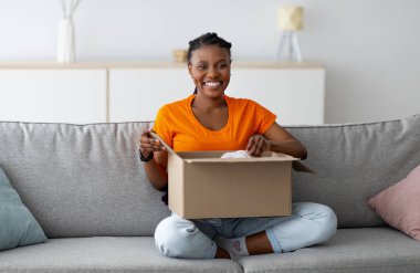 Karton paketi açan neşeli siyahi bir kadın, bacak bacak bacak üstüne atmış koltukta oturuyor, içerde kameraya gülümsüyor. Genç kadın tüketici çevrimiçi market siparişini açıyor, teslimat hizmetinden memnun.