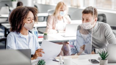 İş yerindeki koruma görevlileri. Koruyucu maskeli Afrikalı Amerikalı kadın, salgın sırasında ofis içi koruyucu camlar vasıtasıyla dizüstü bilgisayarları ve antiseptikleri olan adama dokümanları gösteriyor.
