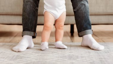 Baba ve küçük bebek birlikte duruyor, sevgi dolu baba, küçük kızının eve ilk adımını atmasına yardım ediyor. Babalar ve Çocuk Bacakları. Sevimli Aile Anları, Babalık Konsepti. Kırpılmış