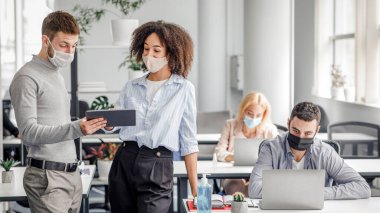 Meslektaşların iş yerindeki hatalar hakkında tartışması. Guy ve koruyucu maskeli Afrikalı Amerikalı kadın, kitle imha silahı salgını sırasında işçilerle birlikte modern ofis içinde tablete bakıyor ve konuşuyorlar.