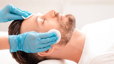 Orta yaşlı yakışıklı adam güzellik kliniğine gidiyor, erkek yüz bakımı konsepti. Tanınmayan spa görevlisinin, tedavi öncesi pamuk pedi kullanarak sakallı adamı tonerle temizleyişinin yan görüntüsü.