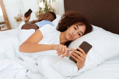 Sosyal sorunlar, alet bağımlılığı ve internet iletişimi. Yoğunlaşmış Afro-Amerikan milenyum bayanı kocasıyla arka arkaya akıllı telefona bakıyor. Modern yatak odasının içinde, fotokopi odası.