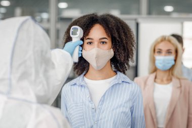 Covid-19 salgını sırasında yeni normaller. Çalışma odasındaki kızılötesi termometreyle koruyucu maskeli çok ırklı işçilerin sıcaklığını kontrol ediyorum.