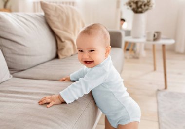 Evdeki oturma odasındaki koltuğun yanında kameraya gülümseyen sevimli bebek. Mutlu Bebek Yalnız Eğleniyor. Çocukluk, Çocuk Bakımı ve Ebeveynlik Konsepti