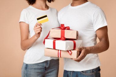 İnternetten hediye sipariş et ve Noel ve yeni yıl için eve teslimat yap. Milenyum Afro-Amerikan erkek ve kadın beyaz tişörtlü. Kutuları ve kredi kartlarını arka planda tutuyorlar.