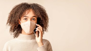 Karantina, sosyal uzaklık ve uzaktan iletişim. Koruyucu maskeli genç Afro-Amerikan bayan telefonda konuşuyor ve hafif krem arka planda izole edilmiş kameraya bakıyor.
