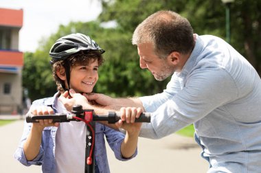 Güvenlik kavramı. Gülümseyen yetişkin babanın portresi parkta küçük çocuğa koruma kaskı takıyor cesur oğluna elektrikli scooter sürmeyi öğretiyor. Hafta sonu etkinliğimiz